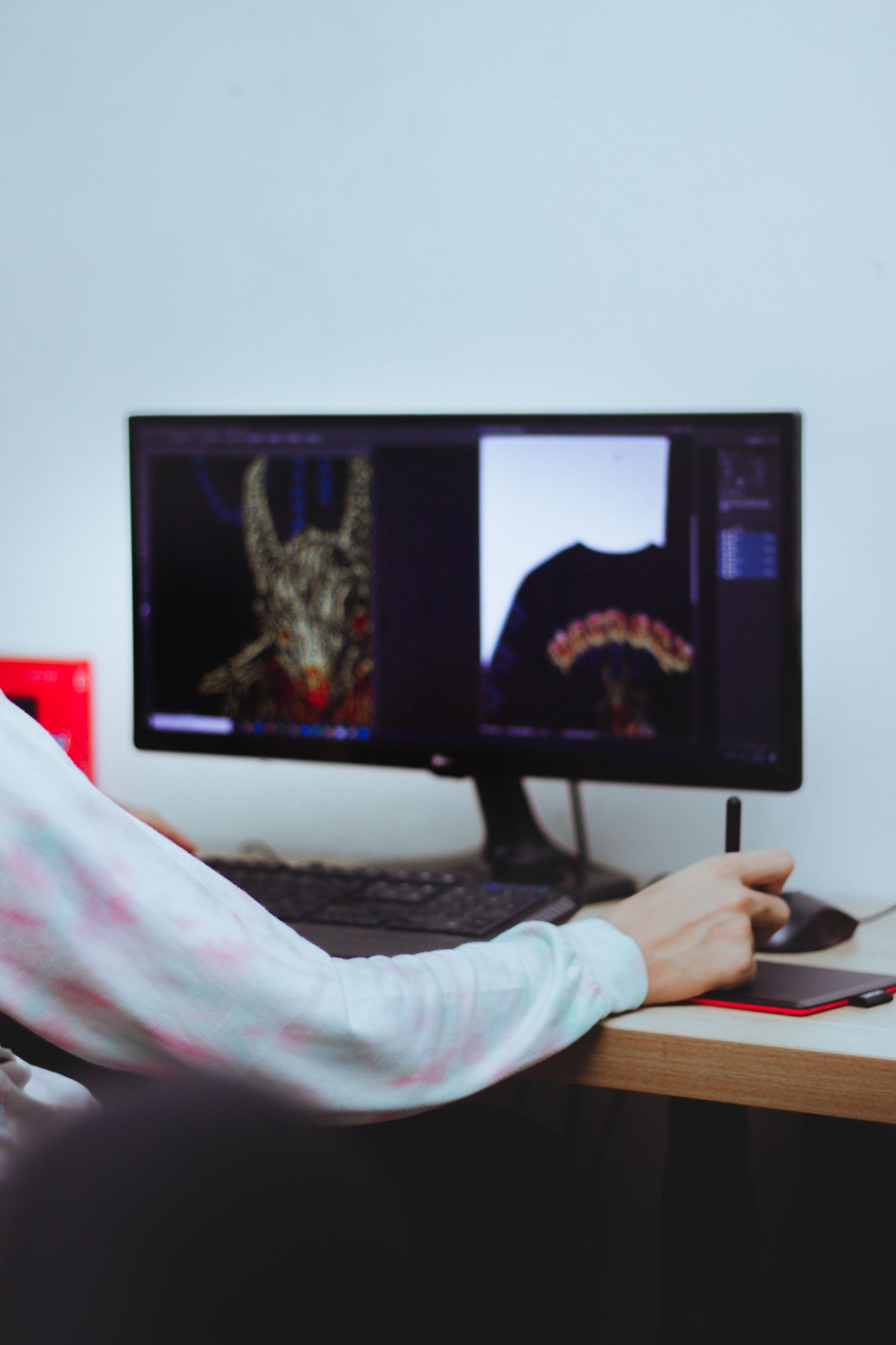 A person seen from behind at a desk, working on an ultrawide monitor with a pen tablet and stylus, editing apparel graphic artwork — a dark illustrated design and a t-shirt mock-up on screen.