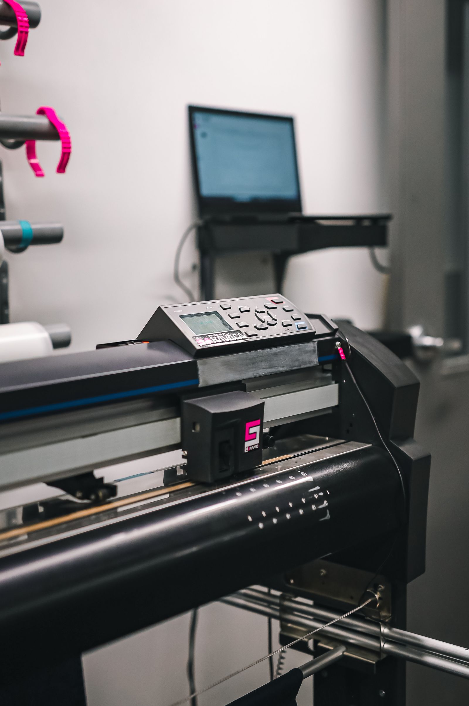 A wide-format vinyl cutter-plotter with a black control panel, media roll-holders with magenta and teal end-caps behind it, and a laptop on a stand in a small print production room.